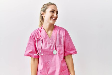 Foto de Mujer rubia joven vistiendo uniforme de enfermera rosa sobre fondo aislado mirando hacia otro lado con sonrisa en la cara, expresión natural. riendo confiado. - Imagen libre de derechos
