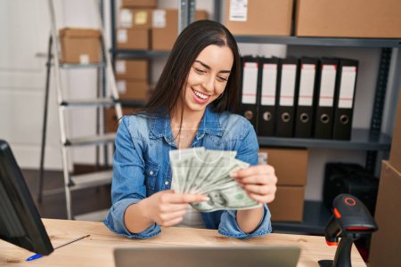 Young beautiful hispanic woman ecommerce business worker using laptop holding dollars at office