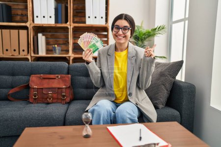 Photo for Young hispanic woman working at consultation office holding money smiling happy pointing with hand and finger to the side - Royalty Free Image