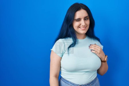 Foto de Young modern girl with blue hair standing over blue background winking looking at the camera with sexy expression, cheerful and happy face. - Imagen libre de derechos