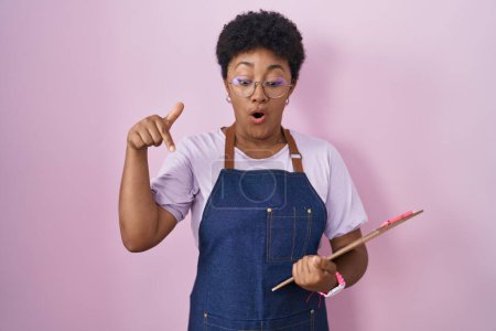 Foto de Mujer afroamericana joven con delantal de camarera profesional sujetando el portapapeles apuntando hacia abajo con los dedos mostrando publicidad, cara sorprendida y boca abierta - Imagen libre de derechos