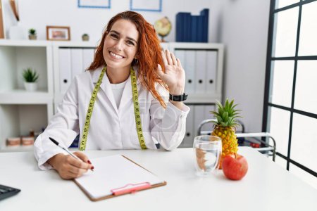 Téléchargez les photos : Jeune femme rousse nutritionniste médecin à la clinique renonçant à dire bonjour heureux et souriant, geste de bienvenue amical - en image libre de droit