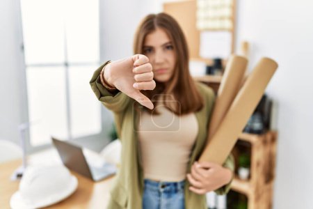 Photo for Young brunette woman holding paper blueprints at the office with angry face, negative sign showing dislike with thumbs down, rejection concept - Royalty Free Image