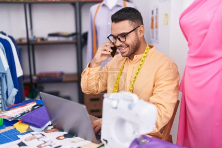 Téléchargez les photos : Jeune homme hispanique tailleur parler sur smartphone en utilisant un ordinateur portable au magasin de tailleur - en image libre de droit