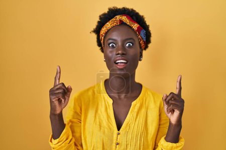 Photo for African young woman wearing african turban amazed and surprised looking up and pointing with fingers and raised arms. - Royalty Free Image