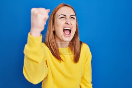 Photo for Young woman standing over blue background angry and mad raising fist frustrated and furious while shouting with anger. rage and aggressive concept. - Royalty Free Image