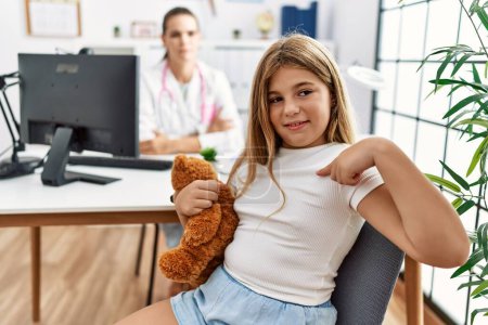 Téléchargez les photos : Blonde little girl at pediatrician clinic with female doctor pointing finger to one self smiling happy and proud - en image libre de droit