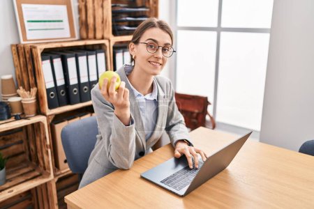 Foto de Young caucasian woman business worker using laptop eating apple at office - Imagen libre de derechos