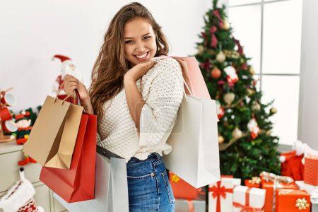 Téléchargez les photos : Young beautiful hispanic woman holding shopping bags standing by christmas tree at home - en image libre de droit