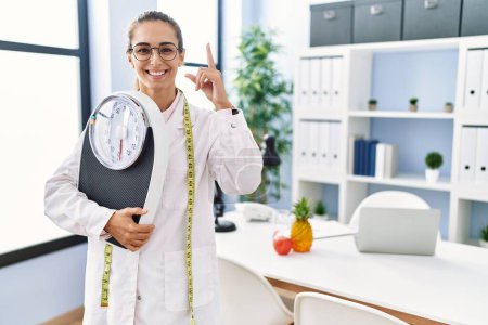 Téléchargez les photos : Jeune femme hispanique comme médecin nutritionniste tenant la machine de pesage surpris par une idée ou une question pointant du doigt avec un visage heureux, numéro un - en image libre de droit