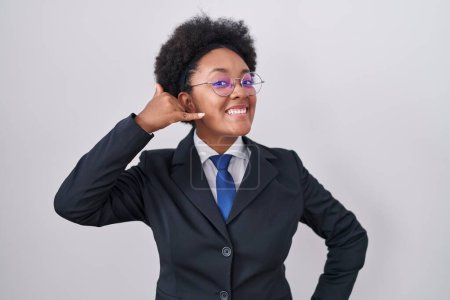 Téléchargez les photos : Belle femme africaine aux cheveux bouclés portant une veste d'affaires et des lunettes souriantes faisant geste téléphonique avec la main et les doigts comme parler au téléphone. communiquer les concepts. - en image libre de droit