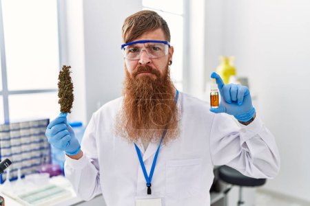 Foto de Redhead man with long beard working at scientist laboratory holding weed and cbd oil skeptic and nervous, frowning upset because of problem. negative person. - Imagen libre de derechos