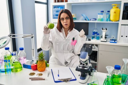 Foto de Mujer hispana joven trabajando en laboratorio científico sosteniendo fruta asustada y sorprendida con la boca abierta para sorpresa, cara de incredulidad - Imagen libre de derechos