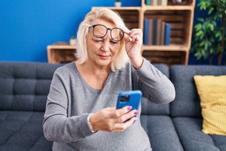 Téléchargez les photos : Moyen-âge femme blonde en utilisant smartphone assis sur le canapé à la maison - en image libre de droit
