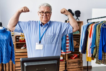 Foto de Hombre mayor con el pelo gris trabajando como gerente en la boutique al por menor mostrando los músculos de los brazos sonriendo orgulloso. concepto de fitness. - Imagen libre de derechos