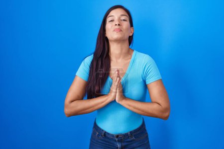 Téléchargez les photos : Jeune femme hispanique debout sur fond bleu mendier et prier avec les mains ainsi que l'expression d'espoir sur le visage très émotionnel et inquiet. mendier. - en image libre de droit