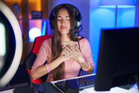 Photo for Young hispanic woman playing video games smiling with hands on chest with closed eyes and grateful gesture on face. health concept. - Royalty Free Image
