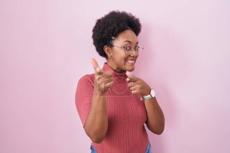 Foto de Hermosa mujer africana con el pelo rizado de pie sobre el fondo rosa señalando los dedos a la cámara con la cara feliz y divertida. buena energía y vibraciones. - Imagen libre de derechos