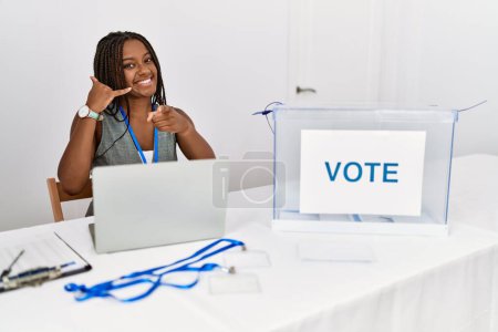Téléchargez les photos : Jeune femme afro-américaine travaillant aux élections politiques assis par bulletin de vote souriant faire parler au téléphone geste et vous pointant du doigt. Appelle-moi. - en image libre de droit