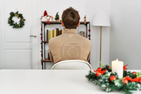 Foto de Joven hombre guapo con barba sentado en la mesa por la decoración de Navidad de pie hacia atrás mirando hacia otro lado con los brazos cruzados - Imagen libre de derechos