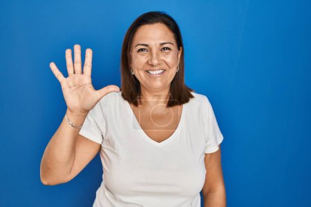 Foto de Mujer madura hispana de pie sobre fondo azul mostrando y señalando hacia arriba con los dedos número cinco mientras sonríe confiado y feliz. - Imagen libre de derechos