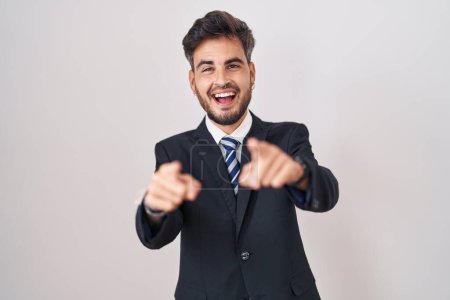 Téléchargez les photos : Jeune homme hispanique avec des tatouages en costume d'affaires et cravate pointant vers vous et la caméra avec les doigts, souriant positif et joyeux - en image libre de droit