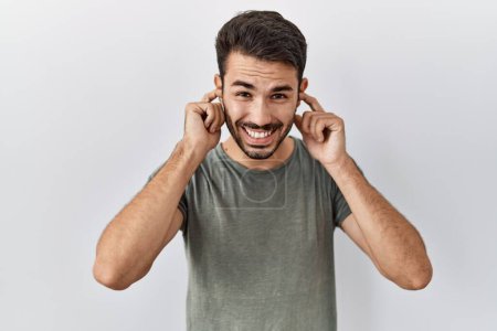 Foto de Young hispanic man with beard wearing casual t shirt over white background covering ears with fingers with annoyed expression for the noise of loud music. deaf concept. - Imagen libre de derechos