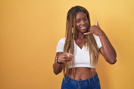 Photo for African american woman with braided hair standing over yellow background smiling doing talking on the telephone gesture and pointing to you. call me. - Royalty Free Image