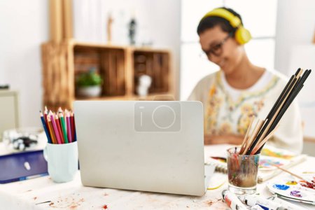 Junge hispanische Frau hört Musik mit Laptop und zeichnet auf Notizbuch im Kunststudio