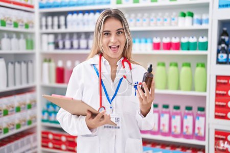 Téléchargez les photos : Young blonde woman working at pharmacy drugstore holding bottle celebrating crazy and amazed for success with open eyes screaming excited. - en image libre de droit