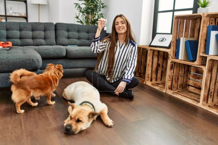 Foto de Joven mujer hispana sosteniendo la llave de una casa nueva sentada en el suelo con perros en casa - Imagen libre de derechos