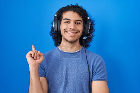 Téléchargez les photos : Homme hispanique aux cheveux bouclés écoutant de la musique à l'aide d'écouteurs avec un grand sourire sur le visage, pointant la main et le doigt vers le côté en regardant la caméra. - en image libre de droit