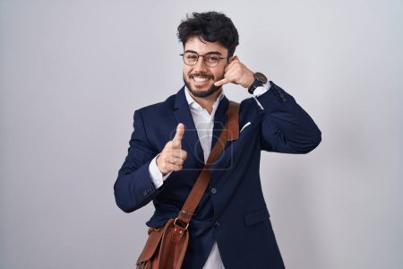 Téléchargez les photos : Homme hispanique avec barbe portant des vêtements de travail souriant faire parler au téléphone geste et vous pointant du doigt. Appelle-moi. - en image libre de droit