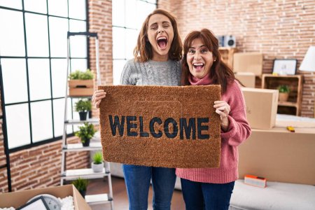 Téléchargez les photos : Mother and daughter holding welcome doormat smiling and laughing hard out loud because funny crazy joke. - en image libre de droit