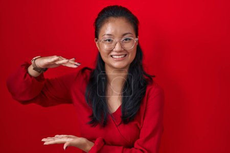 Photo for Asian young woman standing over red background gesturing with hands showing big and large size sign, measure symbol. smiling looking at the camera. measuring concept. - Royalty Free Image