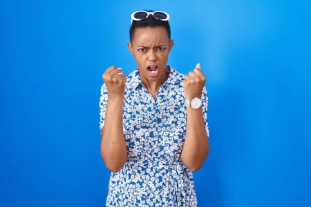 Téléchargez les photos : Femme afro-américaine debout sur fond bleu en colère et fou levant les poings frustrés et furieux tout en criant de colère. rage et concept agressif. - en image libre de droit