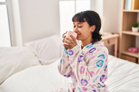 Foto de Joven mujer hispana hermosa bebiendo taza de café sentado en la cama en el dormitorio - Imagen libre de derechos