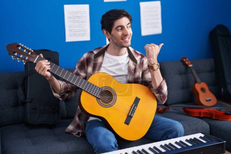 Foto de Joven hispano tocando la guitarra clásica en un estudio de música apuntando con el pulgar hacia un lado sonriendo feliz con la boca abierta - Imagen libre de derechos
