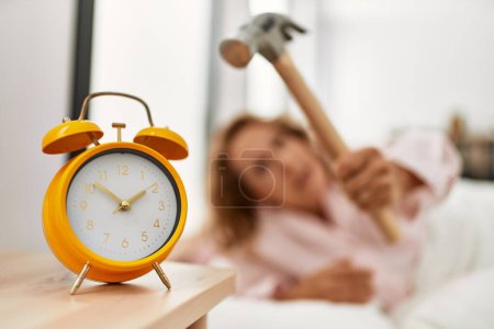 Foto de Mujer caucásica de mediana edad apagar el reloj despertador utilizando martillo acostado en la cama en el dormitorio. - Imagen libre de derechos