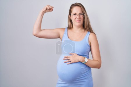 Foto de Mujer embarazada joven de pie sobre fondo blanco fuerte persona que muestra el músculo del brazo, confiado y orgulloso de poder - Imagen libre de derechos