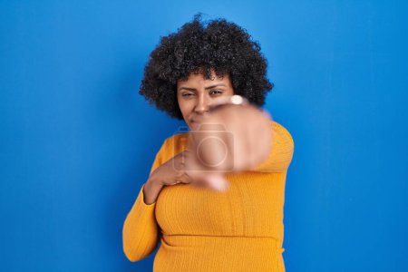 Foto de Mujer negra con el pelo rizado de pie sobre fondo azul puñetazo puño para luchar, ataque agresivo y enojado, amenaza y violencia - Imagen libre de derechos