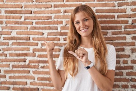 Foto de Young caucasian woman standing over bricks wall pointing to the back behind with hand and thumbs up, smiling confident - Imagen libre de derechos