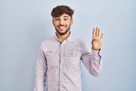 Téléchargez les photos : Homme arabe avec barbe debout sur fond bleu montrant et pointant vers le haut avec les doigts numéro quatre tout en souriant confiant et heureux. - en image libre de droit