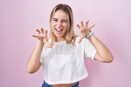 Téléchargez les photos : Jeune femme blonde debout sur fond rose souriant drôle faisant griffe geste comme chat, expression agressive et sexy - en image libre de droit
