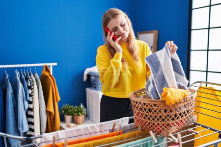 Téléchargez les photos : Young blonde woman talking on smartphone hanging clothes on clothesline at laundry room - en image libre de droit