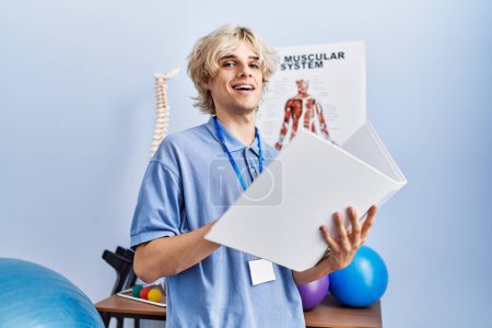 Téléchargez les photos : Jeune homme blond pysiothérapeute souriant document de lecture confiant à la clinique de réadaptation - en image libre de droit