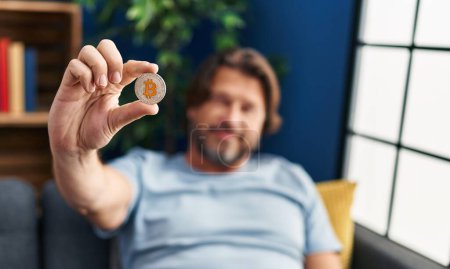 Foto de Hombre de mediana edad sosteniendo bitcoin sentado en el sofá en casa - Imagen libre de derechos