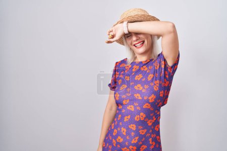 Foto de Mujer caucásica joven con vestido de flores y sombrero de verano que cubre los ojos con el brazo sonriente alegre y divertido. concepto ciego. - Imagen libre de derechos