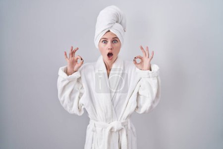 Photo for Blonde caucasian woman wearing bathrobe looking surprised and shocked doing ok approval symbol with fingers. crazy expression - Royalty Free Image