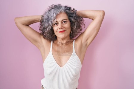 Foto de Mujer de mediana edad con el pelo gris de pie sobre fondo rosa relajante y estiramiento, brazos y manos detrás de la cabeza y el cuello sonriendo feliz - Imagen libre de derechos